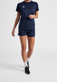 Vrouw in marineblauwe sportkleding, met witte sokken en zwarte hardloopschoenen, staand tegen een effen lichte achtergrond.