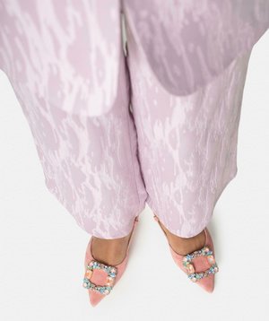 Voeten met roze schoenen met puntige neus en meerkleurige met juwelen versierde gespen, gecombineerd met lichtroze gestructureerde wijde broekspijpen.