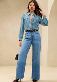 Lichtblauw, gestructureerd blouse met verticale strepen, lange mouwen en twee voorzakken. Hoge taille, wijde blauwe jeans met een bruine riem.