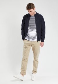 Veste bomber zippée marine par-dessus un T-shirt rayé blanc et marine, assortie à un pantalon beige et des baskets blanches. Tissu lisse, coupe décontractée.