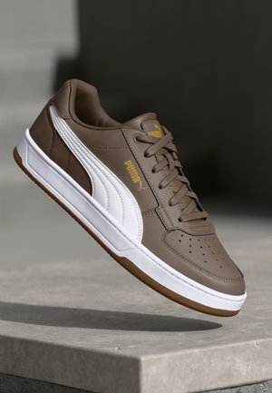 Baskets Puma marron avec une bande blanche, un logo doré et une semelle blanche flottant au-dessus d'une surface en béton sur un fond sombre.