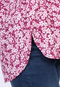 Floral patterned top in pink and white fabric with a curved hem and side slits. Worn over dark denim jeans.