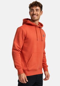 Sudadera naranja hecha de tela suave, que cuenta con un bolsillo frontal, capucha con cordón y un pequeño logo en el pecho. El modelo está posando.