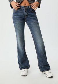 Vyššie vyrezané rozšírené džínsy v tmavomodrom denim, s ošúchaným vzhľadom, kontrastnými švami a surovým lemom. V kombinácii s bielymi teniskami.