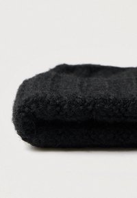 Salocīta melna adīta vilnas šalle ar redzamu tekstūru uz gaiši pelēkas virsmas.