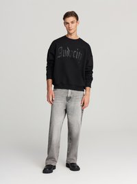 Felpa nera con la scritta "Audacity" in lettere tonali, abbinata a jeans grigio chiaro e scarpe nere chunky. Tessuto liscio, vestibilità comoda.