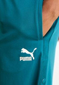 Teal treningsskjorte laget av glatt stoff, med hvit Puma-logo, korte ermer og knappedetalj på siden.