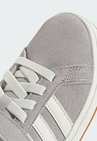 Sneaker en daim gris avec des rayures blanches en cuir, une zone de lacets texturée et un design de lacets plats. La semelle est de couleur marron clair.