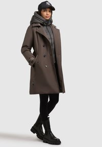 Trench coat impermeabile marrone con bottoni doppi, giacca imbottita grigia sotto, leggings neri aderenti e stivali neri chunky alla caviglia.