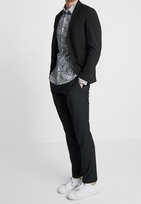 Blazer noir sur une chemise à motif blanche et noire, associé à un pantalon noir et des baskets blanches. Tissu lisse, coupe ajustée.