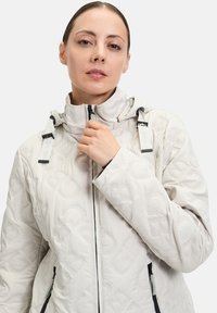 Lys grå quilted jakke med høj krave, der har et mønstret tekstur, lynlåslommer og justerbare snore på hætten.