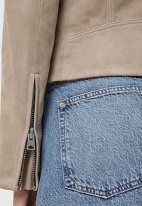 Beige suède jack met een zijrits. Het jack is gecombineerd met blauwe denim, waarbij een achterzak met zichtbare stikseldetails te zien is.