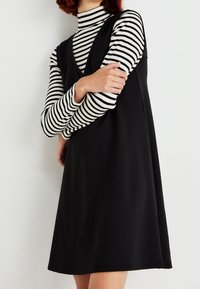 Robe noire sans manches avec un profond décolleté en V, portée sur un haut à manches longues rayé noir et blanc, présentant une texture lisse et une coupe ample.