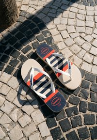 Paire de tongs Havaianas rayées blanc, marine et rouge sur un pavé noir et blanc à motif, la lumière du soleil projetant des ombres.