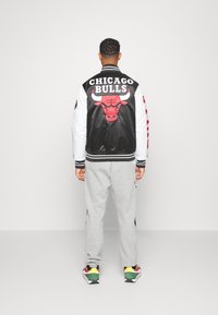 Chaqueta bomber en blanco y negro con el logo de los Chicago Bulls en la parte posterior, combinada con pantalones de chándal grises y zapatillas multicolores.