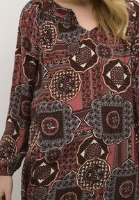 Halenka s dlouhým rukávem v červené a hnědé vzorované tkanině, s propracovanými geometrickými vzory a nařaseným výstřihem.