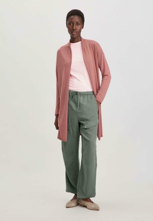 Cardigan lungo e aperto davanti in rosa pallido, indossato sopra una camicia rosa chiaro, abbinato a pantaloni larghi verdi oliva con vita elasticizzata e scarpe beige.