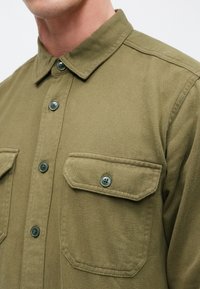 Texturált anyagból készült olívazöld gombos ing, két pipával és gombbal záródó mellzsebbel és klasszikus gallérral.
