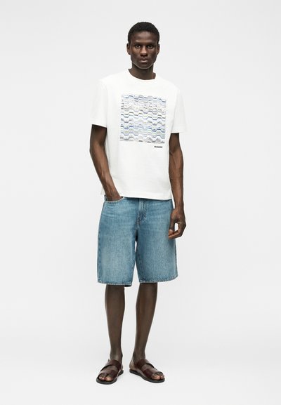 Άνδρας με λευκό κοντομάνικο μπλουζάκι με μπλε κυματιστό μοτίβο, τζιν σορτς μέχρι το γόνατο και καφέ σανδάλια, στέκεται μπροστά σε λευκό φόντο.