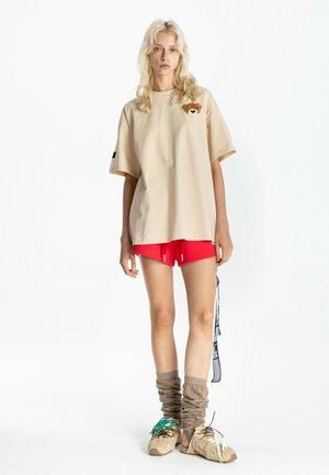 Joven mujer con cabello largo y rubio que lleva una camiseta beige holgada con un pequeño parche de osito, pantalones cortos rojos, calentadores de piernas beige fruncidos y zapatillas beige.