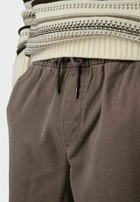 Szare spodnie w twill z elastycznym paskiem, sznurkiem z metalowymi końcówkami i bocznymi kieszeniami, w połączeniu z beżowym i brązowym swetrem w wzory plecionkowe.