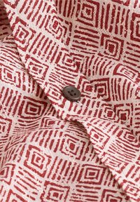 Tissu en coton avec un motif géométrique dans des tons de rouge sur un fond crème. Présente un bouton brun foncé près du pli du tissu.