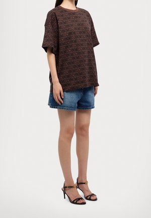 Vrouw draagt een bruin T-shirt met Moschino-logo, blauwe denim shorts en zwarte sandalen met hoge hakken, staand tegen een witte achtergrond.
