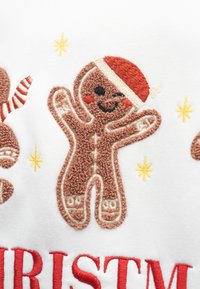 Izšūts piparkūku figūra brūnā krāsā, ar sarkanu Ziemassvētku cepuri un smaidīgu seju, uz baltas fona ar dzeltenām zvaigznēm.