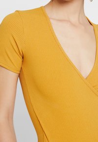 Gul randig bodysuit med djup V-hals och korta ärmar; textilens struktur ger dimension; synliga sömdetaljer längs kanterna.