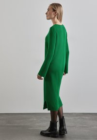 Abito lungo a maniche lunghe verde a coste con uno spacco laterale, abbinato a stivali neri alla caviglia. L'abito ha una vestibilità comoda e un design a girocollo.