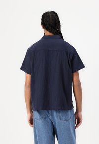 Navy short-sleeve shirt with a textured fabric, classic collar, and straight hem, worn over light blue denim pants.