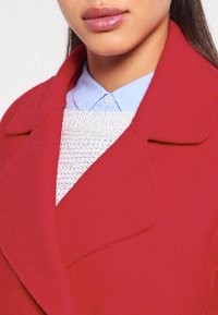 Rode wolmix jas met een notch-kraag, gelaagd over een gestructureerde witte trui en een blauw overhemd, met de nadruk op een gestructureerd design.