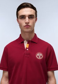 Polo shirt bordeaux con maniche corte, colletto a coste, patta con due bottoni e un accento beige. Presenta un logo circolare sul petto.