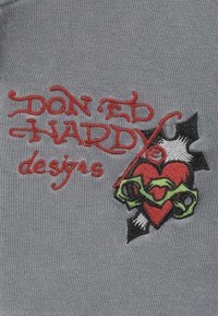 Rood geborduurd "DON ED HARDY designs" tekst met een doorboorde rode hart omringd door groene prikkeldraad op een grijze stof achtergrond.
