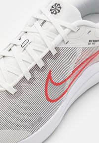 Weißer Laufschuh mit einem Obermaterial aus Mesh, das dünne graue Streifen aufweist, einem roten Nike-Swoosh und weißen Schnürsenkeln. Leichte, strukturierte Sohlengestaltung.
