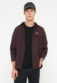 Camicia di flanella a quadri rossi scuri e neri, con bottoni, maniche lunghe, indossata sopra una t-shirt nera. Cappellino nero con logo.