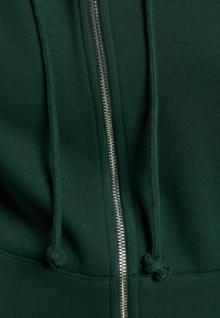 Felpa verde con zip dotata di cerniera metallica, coulisse e texture del tessuto liscia. Sottolinea il comfort e il design informale.