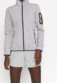 Persoon in een grijze fleece jack met ritssluiting en neongele ritsen, en lichtgrijze shorts met witte tekst op het linkerbeen.
