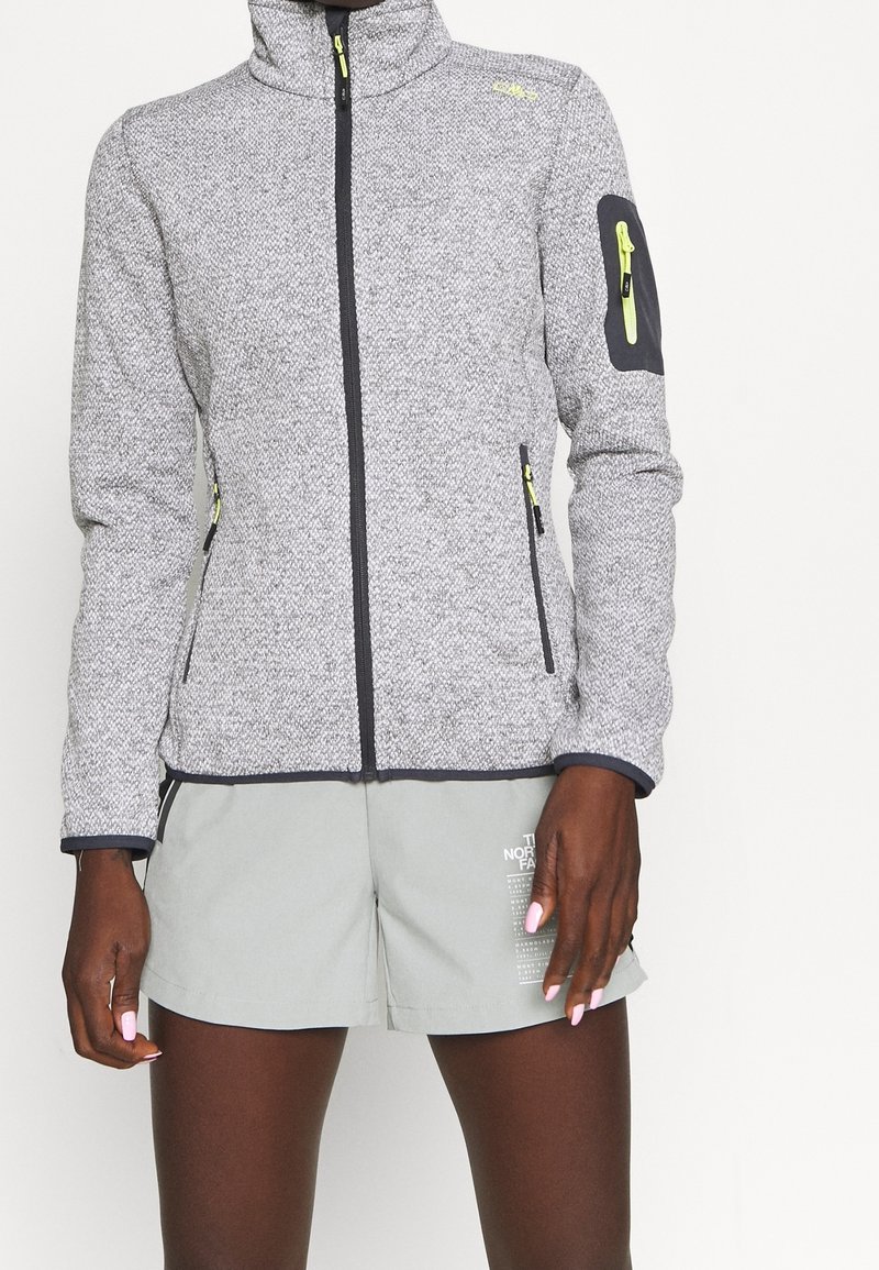 Persoon in een grijze fleece jack met ritssluiting en neongele ritsen, en lichtgrijze shorts met witte tekst op het linkerbeen.