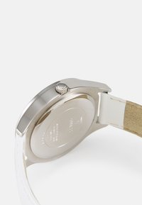 Runder Gehäuse aus silbernem Edelstahl mit strukturiertem Kranz, ausgestattet mit einem weißen Lederarmband und beigem Wildleder auf der Rückseite für zusätzlichen Komfort.