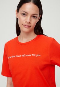 Rode katoenen T-shirt met korte mouwen, voorzien van witte tekst die over de borst leest: "je ware hart zal je nooit in de steek laten."