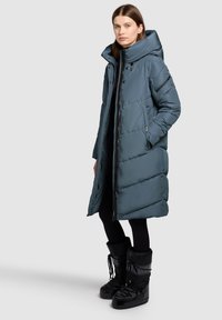 Langer türkisfarbener Steppmantel mit hohem Kragen, versehen mit Reißverschluss vorne und Seitentaschen. Getragen über schwarzen Leggings und schwarzen gefütterten Stiefeln.