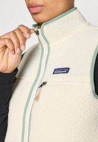Fleecedžep jakna u krem boji s teksturiranim završetkom, mint zelenim obrubom, visokim ovratnikom i prednjim zatvaračem na rajsferšlus. Sadrži patch s logotipom Patagonije.