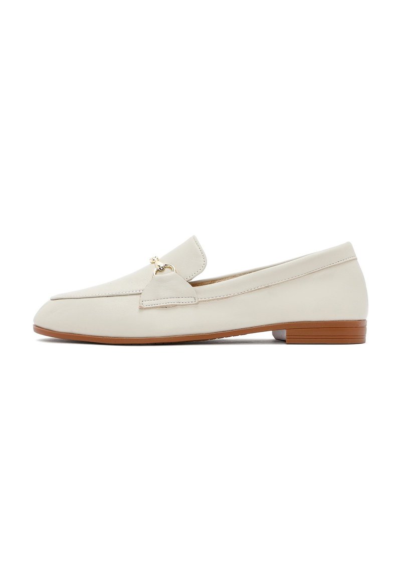 Crème-kleurige leren loafers met een platte, bruine zool. Kenmerkt zich door een voorklep en een kleine gouden accent aan de zijkant. Gladde textuur, minimalistisch ontwerp.