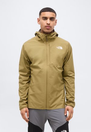 Mann trägt eine olivgrüne The North Face Kapuzenjacke und graue und schwarze Hosen, steht vor einem einfachen hellen Hintergrund.