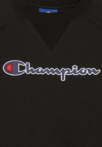Tessuto nero con logo Champion ricamato in lettere bianche e blu contornate, con un emblema "C" rosso e blu sulla sinistra.