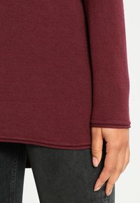 Maglia bordeaux a maniche lunghe realizzata in tessuto morbido, con orlo arrotondato e polsini aderenti, abbinata a pantaloni grigi scuri.