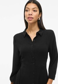 Juodas mygtukų suknelė su apykakle, ilgomis rankovėmis ir prigludusia liemene. Lygus audinys su švelniu blizgesiu ir struktūrizuota siluetu.