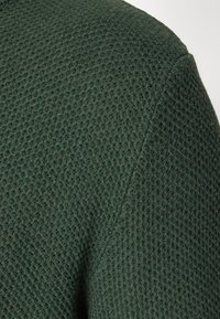 Grüner gestrickter Stoff mit einer strukturierten Waffeloberfläche. Die Oberfläche ist leicht erhöht, was Tiefe und ein weiches, gewebtes Aussehen zeigt.