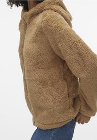 Person som bär en beige, fluffig jacka med huva och långa ärmar, i kombination med blå jeans, stående mot en ren vit bakgrund.